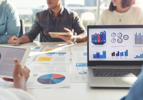 Group of people meeting with technology and paperwork. Close up of digital tablet and laptop computer with market analysis data on the screen. The documents have financial or marketing figures, graphs and charts on them. Multi ethnic group including Caucasian, latin and African American