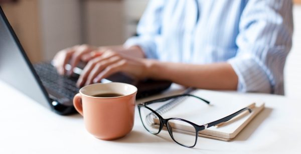 Remote working from home. Freelancer workplace in kitchen with laptop, cup of coffee, spectacles. Concept of distance learning, isolation, female business, shopping online. Close up of woman hands.
